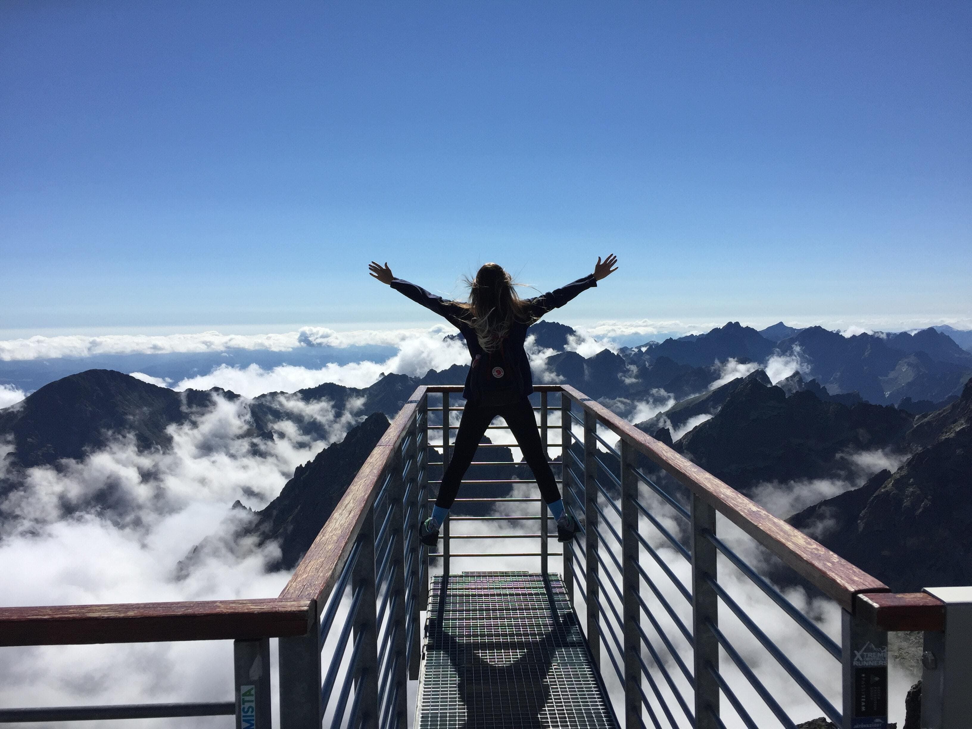 Person standing with arms raised on a viewing platform above mountain peaks and clouds.